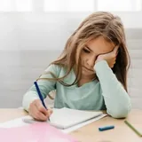 طفلة عند مكتبها تكتب في دفترها وهي تبدو متعبة